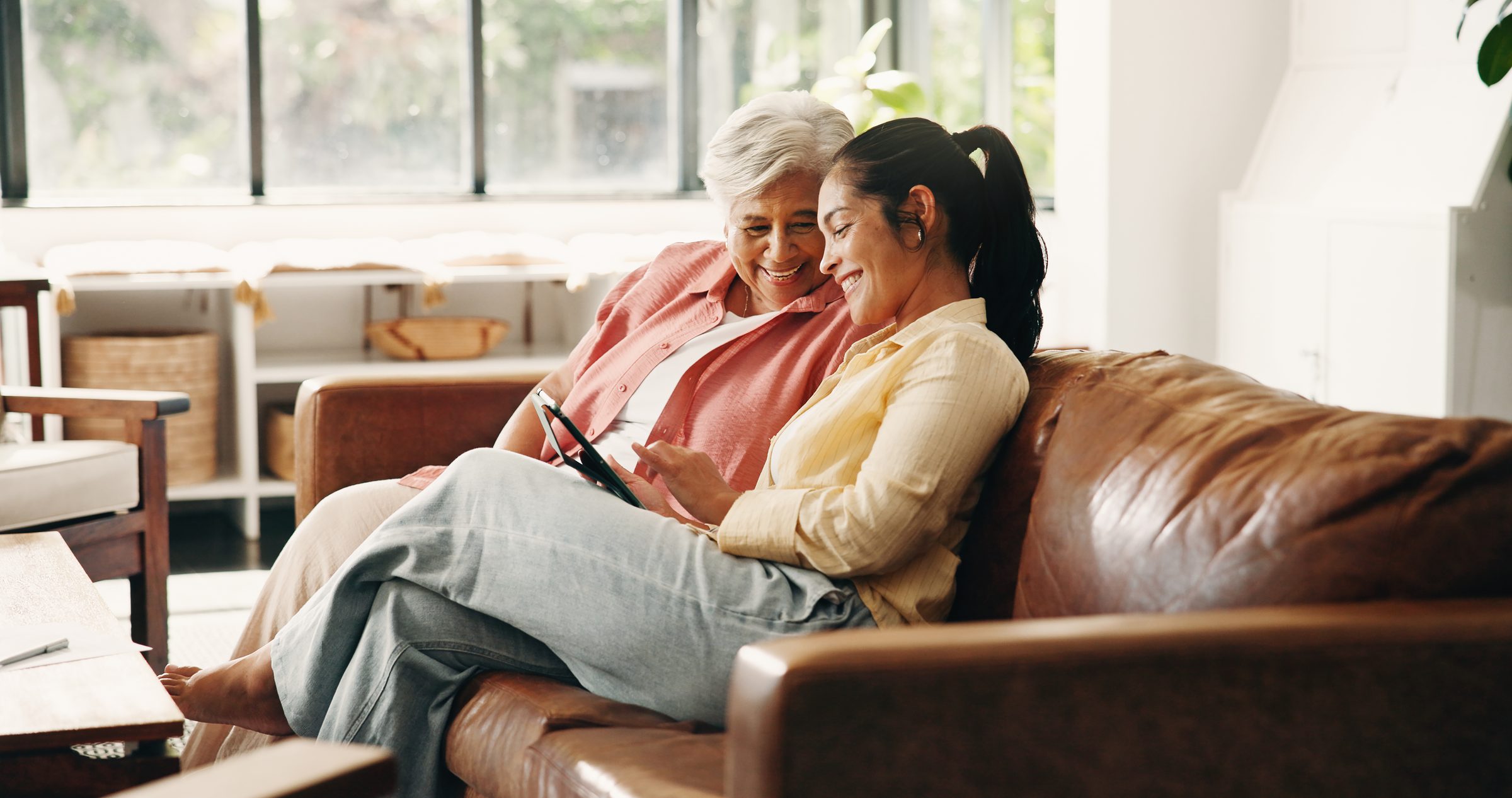 An older woman and her daughter sitting close together on a sofa, looking at a tablet