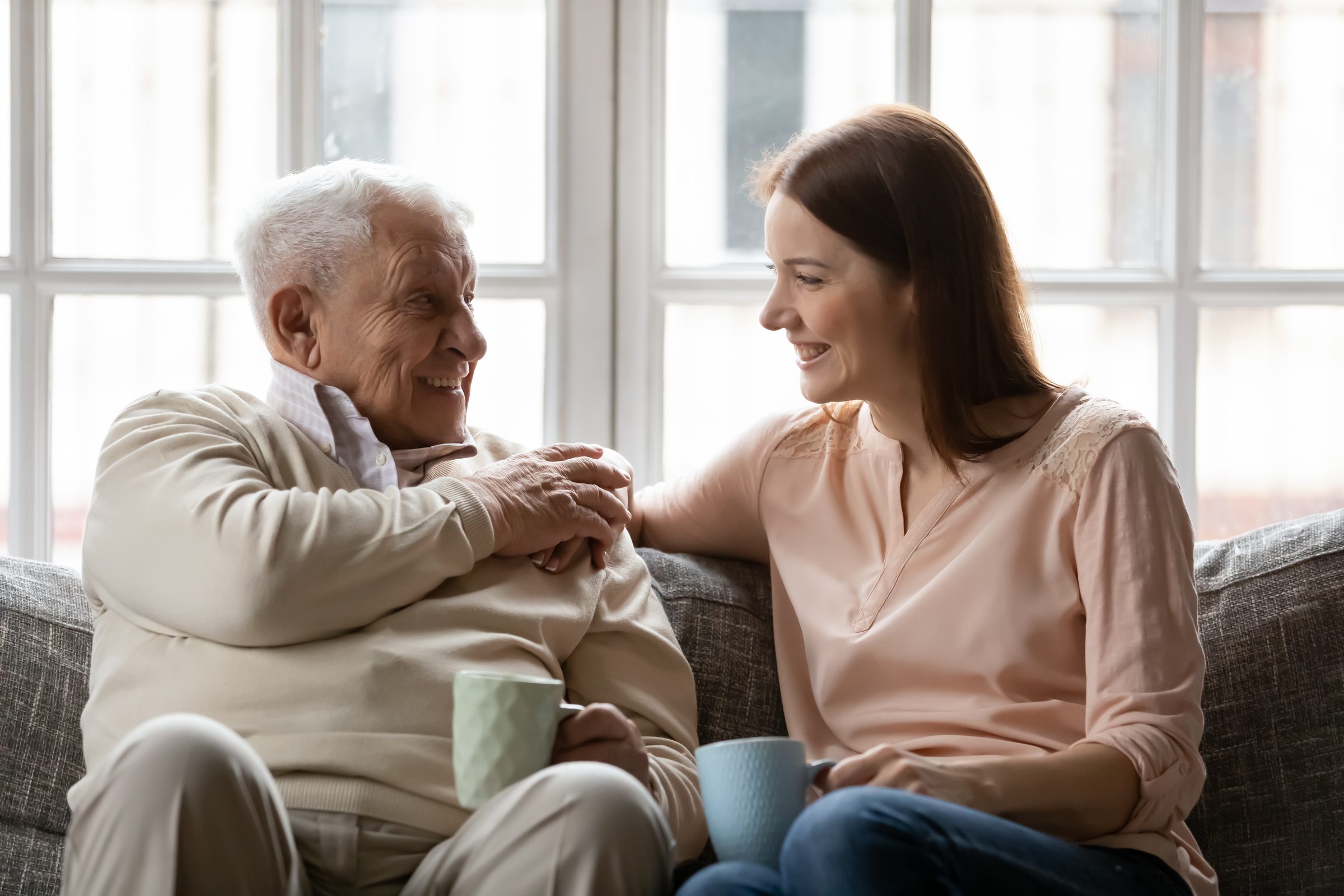 An older man and his adult daughter laughing over coffee on the sofa
